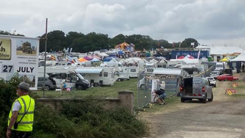 A long distance shot of the show site. There are dozens of campervans and tents parked up to the left. hand side and a red and yellow circus top tent in the distance, as well as  a number of cars parked up along a section of fencing with festoon lights on it. A warden wearing a yellow high vis vest stands to the left of the image near a sign for the show.