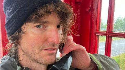 William Arnold wearing a raincoat and hat standing holding a phone in a red phone box.