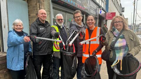 Luisa Scott among a group of five people with litter pickers and bin bags on Gateshead High Street. She has dark hair, tied back, and is wearing an orange hi-vis jacket. They are all looking directly into the camera and smiling.