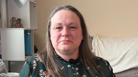 Elaine is looking at the camera. She has long brown/grey hair and is wearing a green floral top with a high neckline.