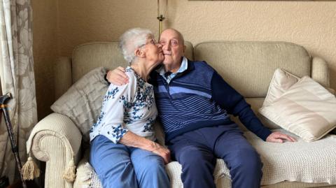 Bob and Pat Page sat on a sofa in a living room. He has his arm around her and she is placing a kiss on his cheek.