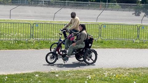 Two individuals ride specially adapted bicycles along a path lined with green grass and safety barriers.