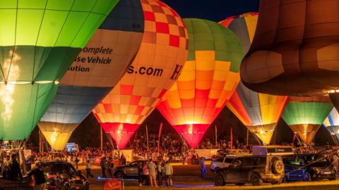 Six hot air balloons are seen tethered on the ground at the Bristol Balloon Fiesta. The picture is taken at night during the nightglow event. There are people and vehicles between the balloons, which are firing their burnings to create light. In the background the crowd on the edge of the arena can be seen
