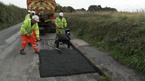 Workers in hi-vis are carrying out work on a road. They appear to be fixing potholes . The four workers are wearing hard hats and are standing in a country lane next to a lorry.