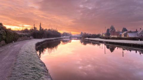 Reflective water with a pink undertone near Sharpness Docks in Gloucestershire. The sun is rising and it looks frosty on the grass surrounding the water. 