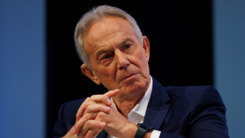 Tony Blair in a navy suit crossing his hands, deep in discussion at a conference