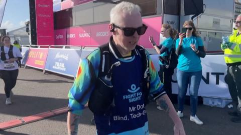 Clarke Reynolds has short white hair. He wears a tie dyed blue, green, red and yellow t shirt and a blue vest over the top which reads Fight for Sight. He has black RayBan sunglasses on.