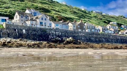 The sea washes into a granite wall with houses behind, a grassed hill rising above and blue sky