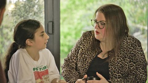 A girl with long dark hair and a white T-shirt with a pink and green print on the front looks towards a woman with brown hair, glasses and wearing a leopard-print jacket. A garden - through glass in the background - is out of focus.