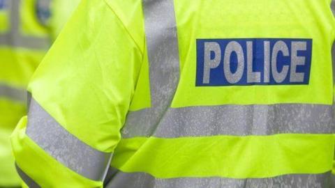 Three police officers with their backs to the camera. They are each wearing a yellow fluorescent jacket which says POLICE on the back in silver writing. 