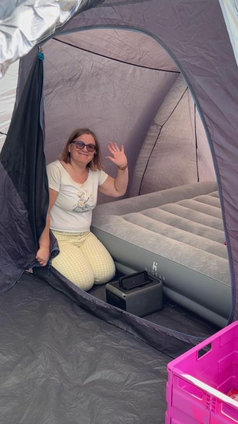 A woman kneeling on the ground inside of a tent pod. A blown up airbed is next to her. The woman is holding her hand in the air as she waves at the camera.