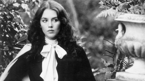 A black and white image of a woman with brunette hair in a garden with stone ornaments. 