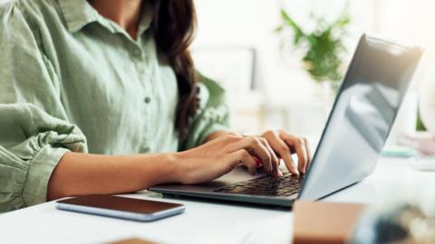 A woman with dark hair wearing a green button-up shirt with puffed three-quarter length sleeves. She is sitting at a white desk, typing on a grey laptop. In the background there is a plant out of focus.