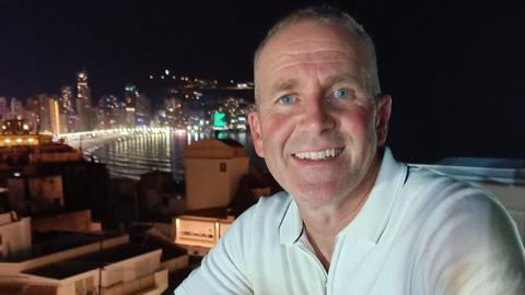 Les Howard, who is wearing a white polo shirt, is starring directly at the camera and is smiling. There is a city skyline in the background. 
