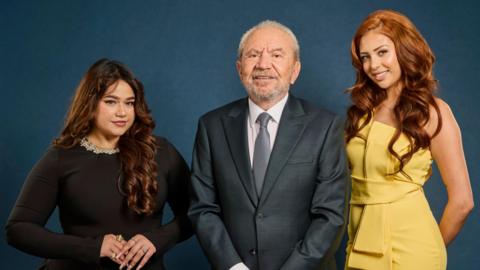 Three people pose for a studio photograph against a dark blue backdrop. Lord Alan Sugar stands in the centre wearing a dark suit and tie. Karishma Vijay stands to one side wearing a black dress, while finalist Pascha Myhill stands on the other side wearing a yellow sleeveless outfit.
