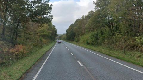 A country road with a black car driving away into the distance. There is thick foliage and trees on both sides of the road.
