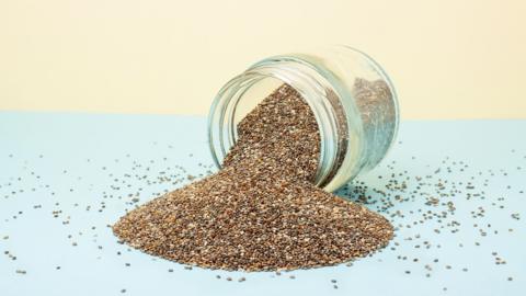 A glass jar on its side, which is full of seeds including chia seeds which are spilling out on a blue and pale yellow background