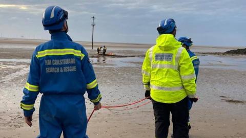 Three members of HM Coastguard Search and Rescue, dressed in yellow and blue high visibility jackets, stand facing the muddy area of Leasowe Bay where a rescue is taking place on the beach.