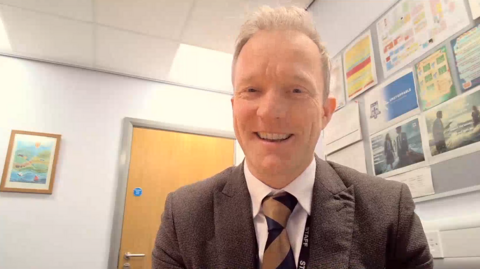 Tony McCabe is a middle-aged man with fair hair. He wears a brown suit, light shirt and stripey tie. He has a lanyard that reads 'staff'. There is a classroom display in the background.