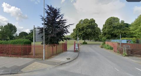 Fenced entrance with sign at school - there are tree-lined paths and glimpses of buildings.