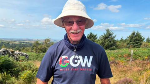 John Parry stands smiling at the camera, with a view of north Pembrokeshire and the Pembrokeshire coast behind him. John is wearing a 'grandad's great walk' fundraiser logo t-shirt, with sunglasses and a sun hat. He wears sunglasses and a white moustache.