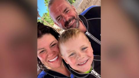 Giselle, a woman with dark hair, Charlie a man with grey hair and a beard and a young Hendrix who has light hair, they all have bright smiles while on holiday wearing wetsuits.