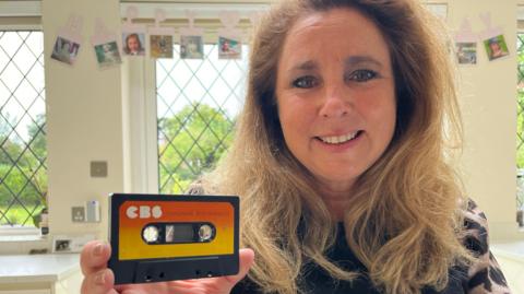 Louise Camby smiles at the camera and holds up an orange and yellow cassette tape on which you can just see the words Dungeons & Dragons