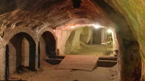 A tourist attraction underground cave in Reigate, with a ramp on the floor and artificial lighting on the walls for visitor access. To the left are two arches, which seem to be made out of brick.