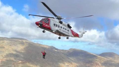 A red and white coastguard helicopter lifting two people in the air on a winch from the mountains in Eryri National Park. The sky is blue with patches of sun hitting the grassy mountain range in the background. 