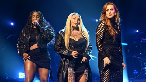 Three woman stood on a stage wearing black. They are holding microphones and singing.