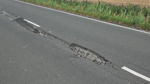 A road on tarmac with a cracked part in the middle is next to a grass verge.