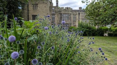 An exterior image of Bolling Hall in Bradford - in the foreground is a green lawn and purple flowers 