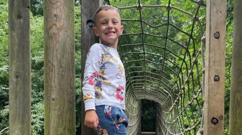 Timotej Borrett, a five-year-old boy standing on wooden climbing frame, in front of a rope-covered tunnel. 