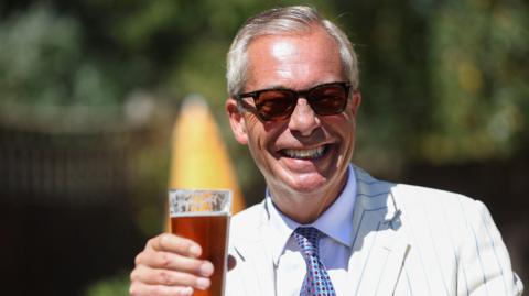 Nigel Farage smiling wearing sunglasses and holding up a pint of beer that is not quite full to the brim.