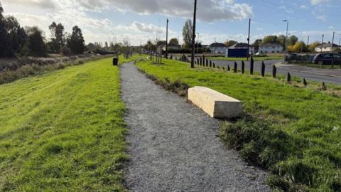 A gravel path surrounded by grass. There is a large stone seat next to the path. It is next to a road, and there are houses on the other side of the road.