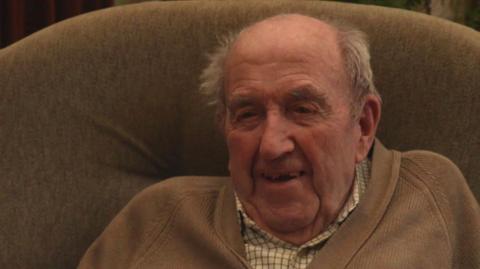 A photo of Alfred 'Barney' Barnett looking at the camera. He is wearing a chequered shirt with a jumper over the top while sitting in a chair.