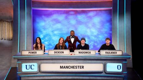 Four students sat at a table. There are name tags on the table (left to right POWER, DICKINSON, MADGWICK, FAULKNER). There is the word Manchester and UC on the front of the table. Behind the four sat down students is the presenter standing up. He is wearing a dark red suit. 