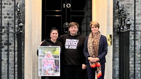 A woman is holding a large picture of an elderly relative next to a man in a black hoodie with the words Regulations and Justice for Jessie written on them. Next to those two is MP Emma Hardy, wearing a blue suit and leopard-print scarf