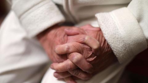 The clasped hands of an elderly woman are resting on her lap in this close up. she is wearing a white fleece top with her cuffs folded over, and white trousers.