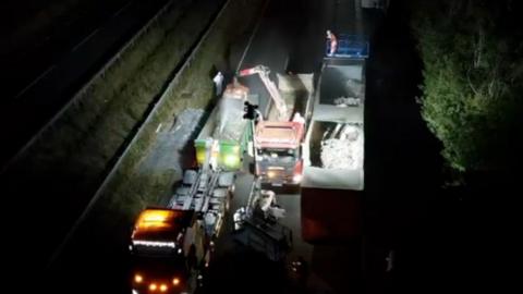 A night-time clean-up operation on a dual carriageway, viewed from an elevated position. A piece of heavy machinery with a lifting arm is positioned alongside the trailer and appears to be removing or handling material from inside it.