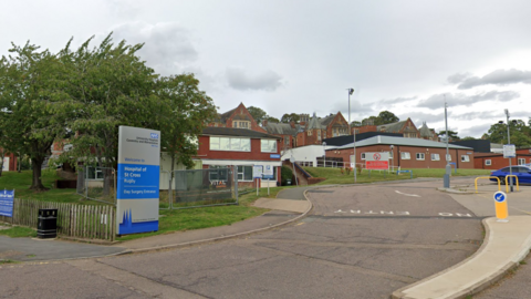 The front of the Hospital of St Cross with a blue and silver NHS sign and brick buildings in the background