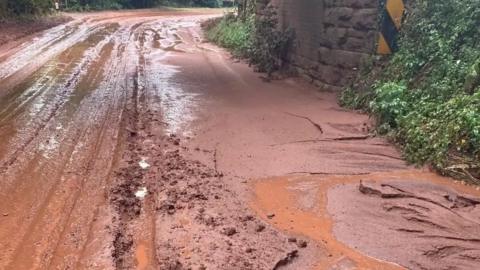 Deep red mud is visible on a country road. Tyre tracks can be seen in the surface. The road is bordered by a stone wall and has green shrubs also alongside it