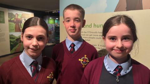 Three children stand together in an exhibition space. They are two girls and a boy all dressed in a school uniform of maroon jumper and pale blue shirt with a striped tie. The trio are standing in front of display boards in a museum. The lighting is quite dark but all three are smiling. 