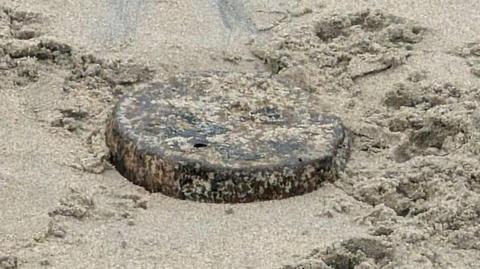 A flat, circular piece of metal can be seen partly buried in the sand. It's rusted, with patches of brown and black and silver flecks underneath.