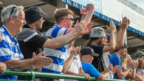 Oxford City fans cheer from the stands, in their blue and white striped shirts.