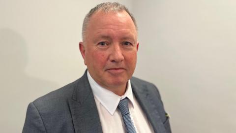 Deputy Liam McKenna - A man with black and grey hair looking towards the camera. He's wearing a blue suit jacket, a white shirt and a blue and white dotted tie. 