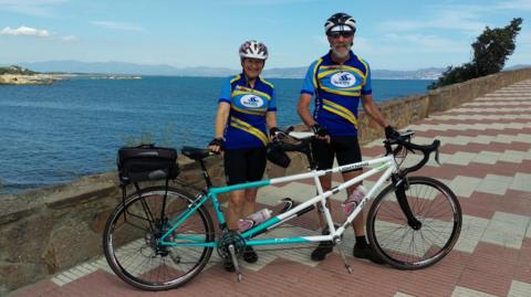 Janet and Barry stood holding a tandem bike in front of them. They are both wearing black cycling shorts, blue and yellow cycling t-shirts, and white helmets. The bike is painted light blue and white. They are stood on pavement in front of the sea. The sky is blue.