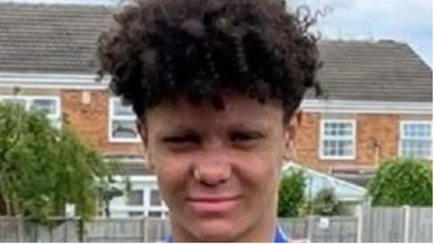 Harleigh has dark curly hair and is wearing a blue top. He stands in front of houses with a fence in front and trees behind them.