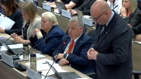 A man with a bald head and glasses, wearing a dark suit, shirt and tie, stands up behind a row of desks to speak during a council meeting. To the side of him sit other men and women at the same desk involved in the meeting. There is also a row of desks with people sat down behind him. Each person has a sign with their name on in front of them. 
