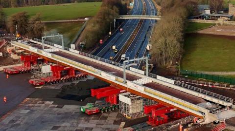 An aerial view of the new bridge being positioned over the M6. It is a light grey with bright red supports underneath rolling it into place.